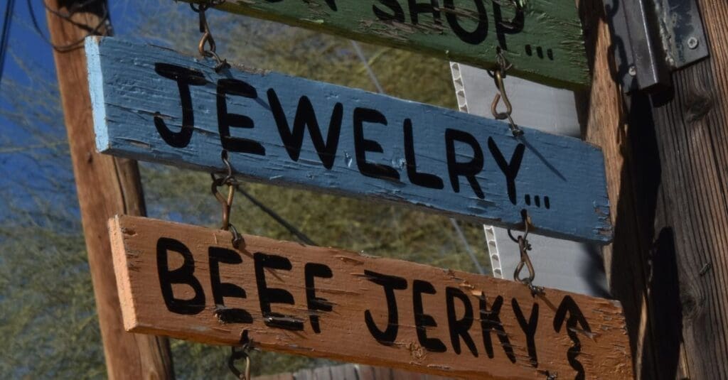 Two rustic wooden signs hanging on chains; blue sign reads 'JEWELRY' and orange-brown sign reads 'BEEF JERKY'.